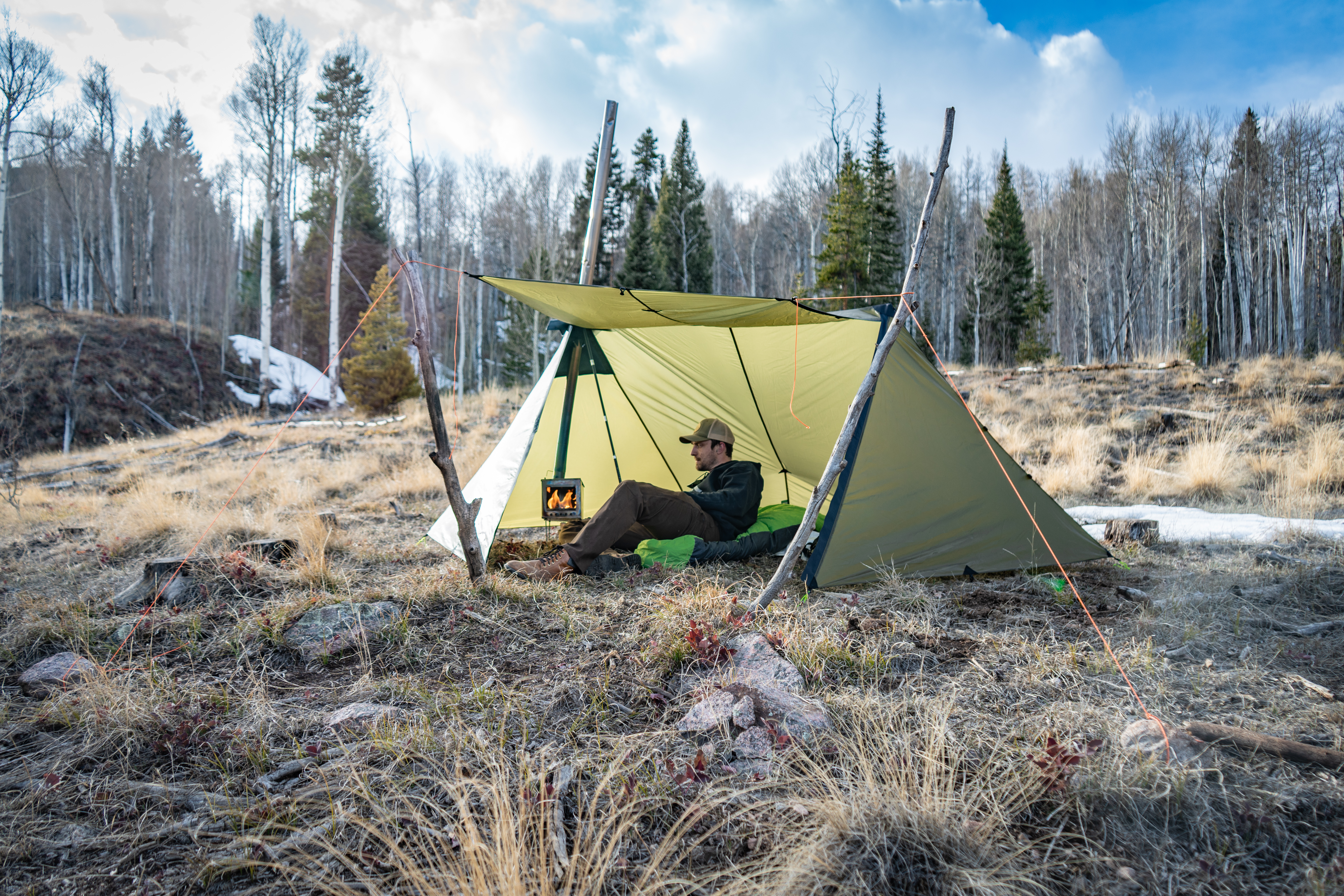 Guardian 2.0 on an alpine hillside with the awning up, a 6 foot tall man alying inside the shelter with a titanium wood stove inside the shelter as smoke belows from the stovepipe
